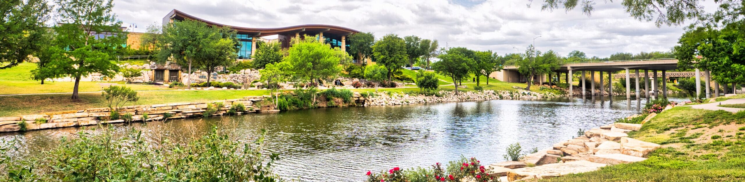 Scenic view of a tranquil riverfront with a park, greenery, and a modern building in San Angelo, Texas.
