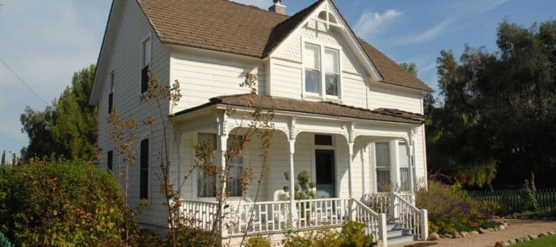 A classic white two-story house with a brown roof and a wraparound porch is surrounded by trees and shrubs under a sunny sky